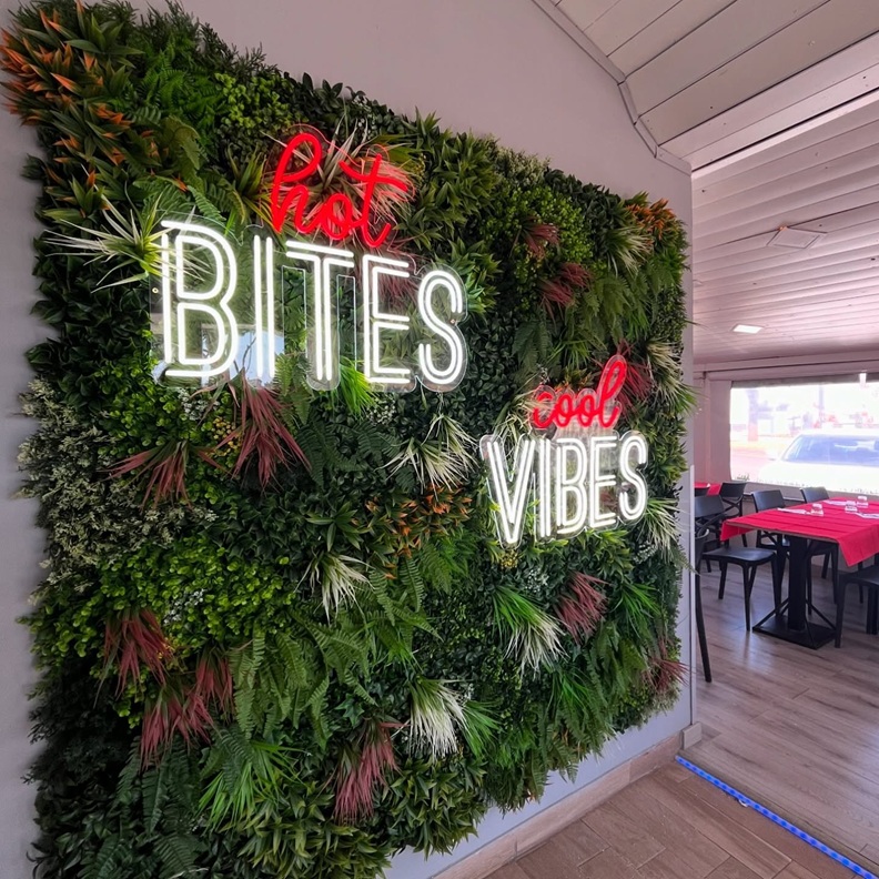 Hot Bites Cool Vibes neon sign on a pizzeria greenery wall