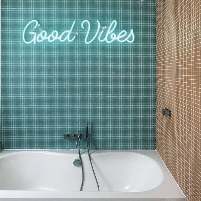White Good Vibes neon sign above a bathtub on a teal tiled wall