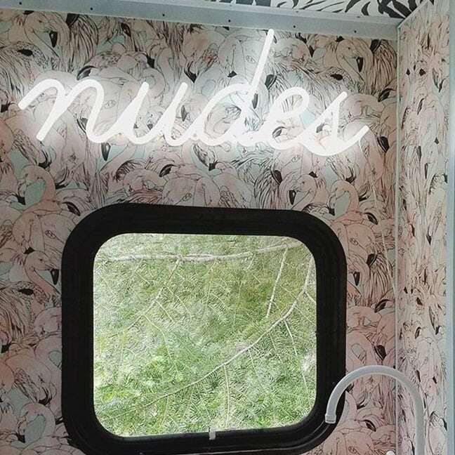 White Nudes neon sign above a bathroom mirror with floral wallpaper
